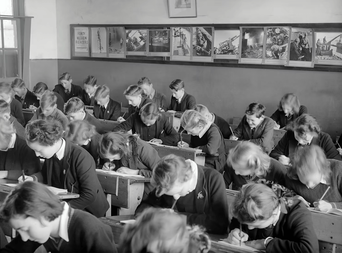 Des élèves en uniforme, penchés sur leur pupitre, passent un examen dans une salle de classe en noir et blanc.