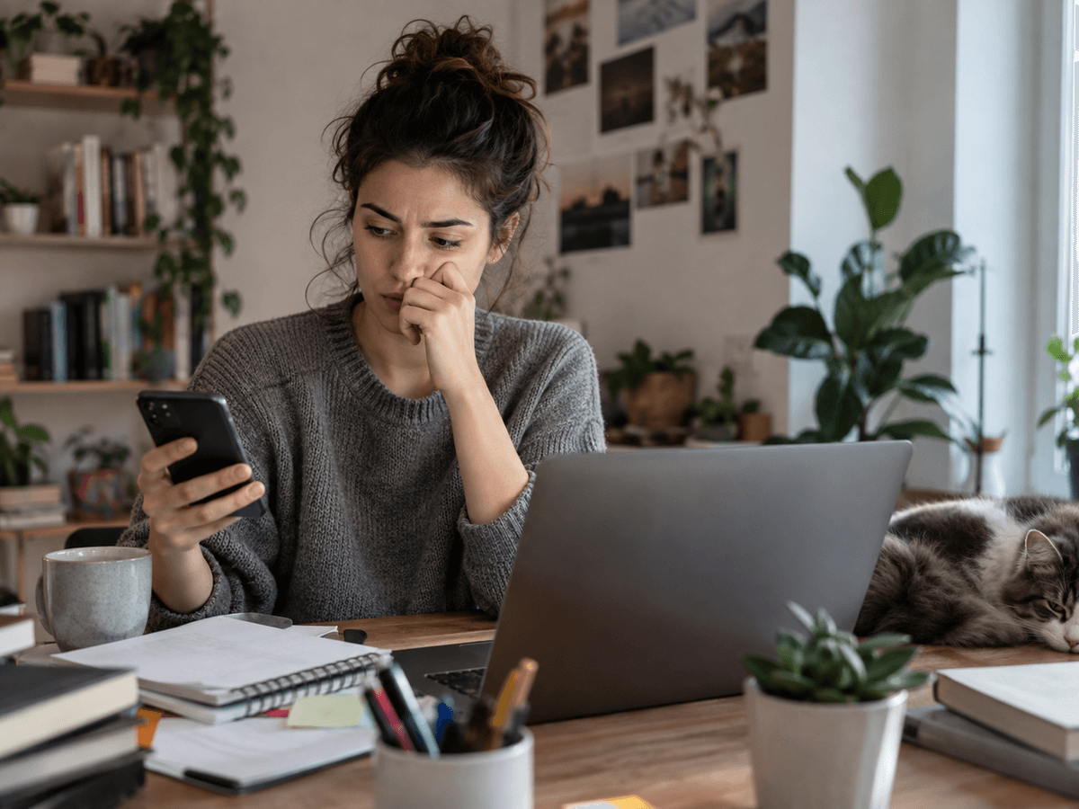Une femme à son bureau a l’air stressée en regardant son téléphone plutôt que son ordinateur portable ouvert.